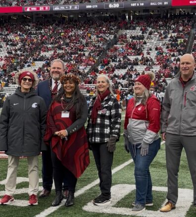 Dr. Gerry Ebalaroza-Tunnell stands on the field at Martin Stadium during a WSU football game, surrounded by university leaders and alumni. She wears a red patterned wrap and a traditional Hawaiian lei headpiece. The group, including WSU President Elizabeth Cantwell (far left) and Chancellor Dave Cillay (far right), poses together smiling as the stadium crowd fills the background.