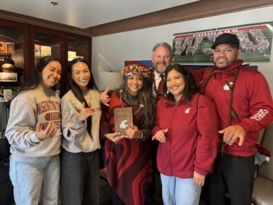 Dr. Gerry Ebalaroza-Tunnell stands with WSU Asian Pacific Islander Desi American (APIDA) student leaders, all smiling and making shaka hand signs. Dr. G holds her Society of Distinguished Alumni award plaque and wears a traditional Hawaiian lei headpiece and a red patterned wrap. The group is gathered indoors in front of WSU memorabilia.