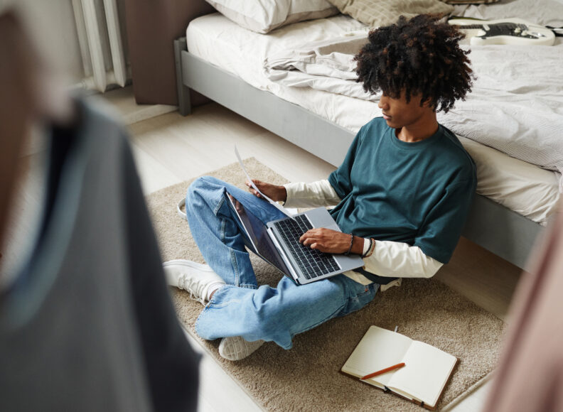 Teenage student sitting on floor at home or in college dorm and studying on a laptop. 