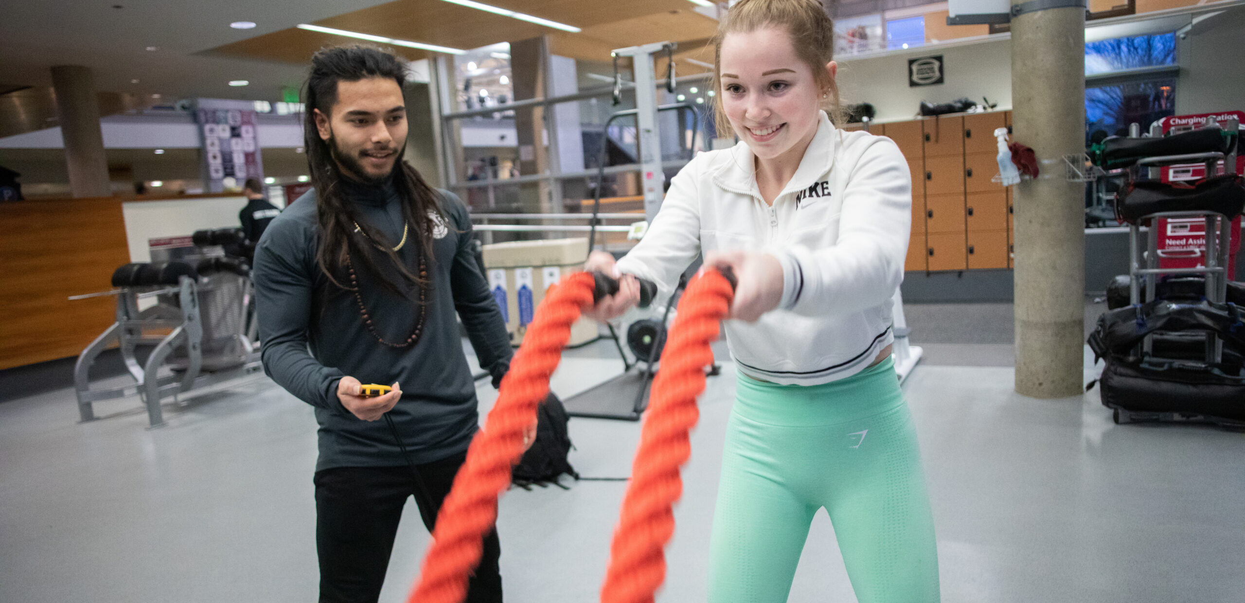 A young woman exercises with battle ropes in a gym while a trainer watches and times her.