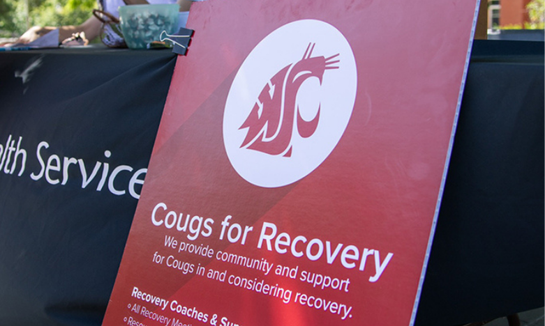 A red WSU Cougars sign on display at a table reads "Cougs for Recovery – We provide community and support for Cougs in and considering recovery." The sign features the WSU Cougar logo and information about recovery coaches and support.