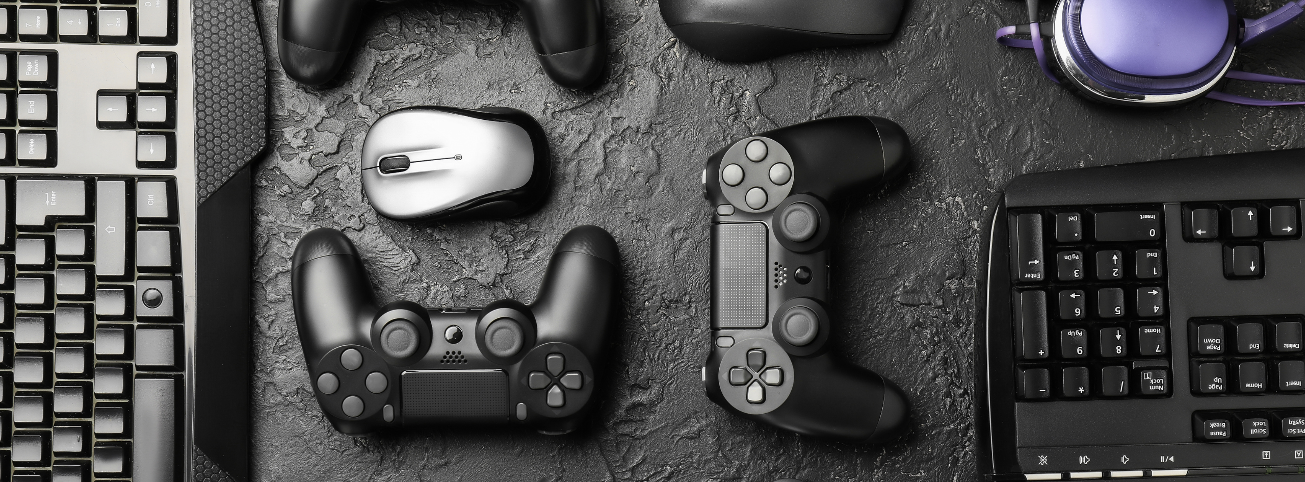 Flat lay of gaming equipment including keyboards, controllers, a mouse, and a purple headset on a dark surface.