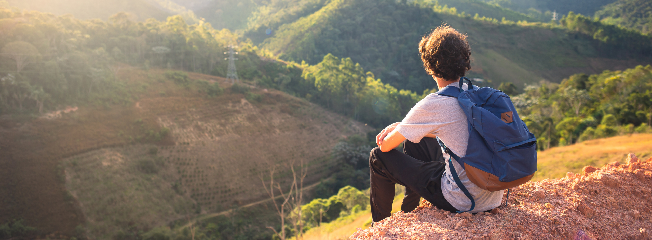 A student with a backpack sits on a hillside overlooking a sunlit valley and forest, symbolizing reflection and personal growth.