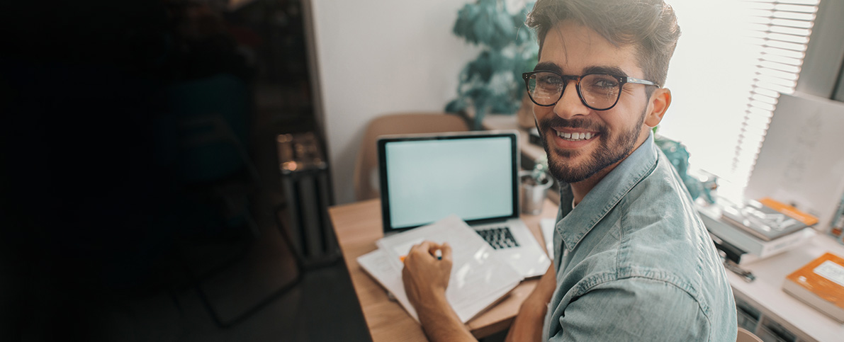 Photo: Student smiles at camera while working at a laptop.