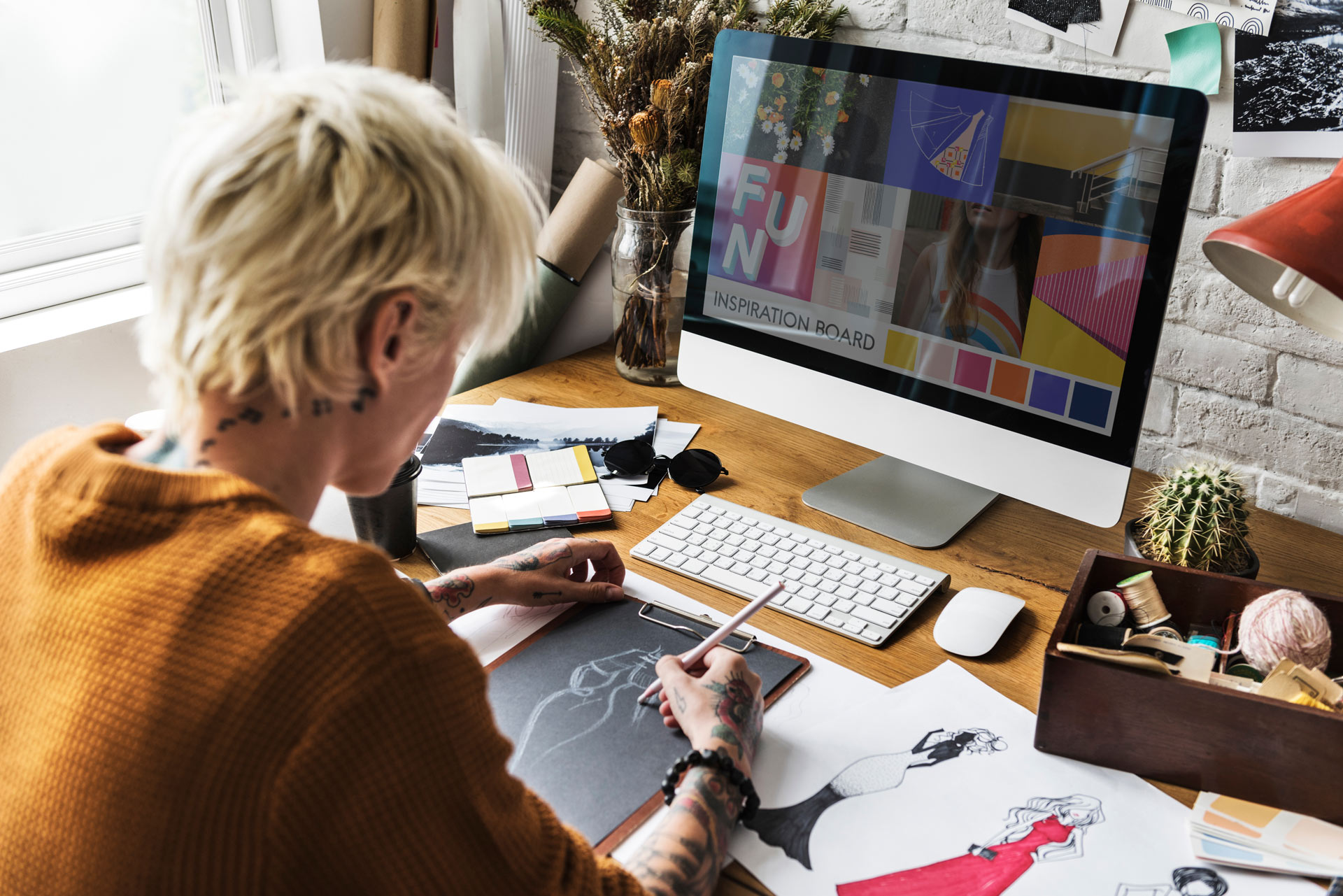 Image of person working with design software as well pencils and paper. Desk is cluttered with drafts and crafts materials.