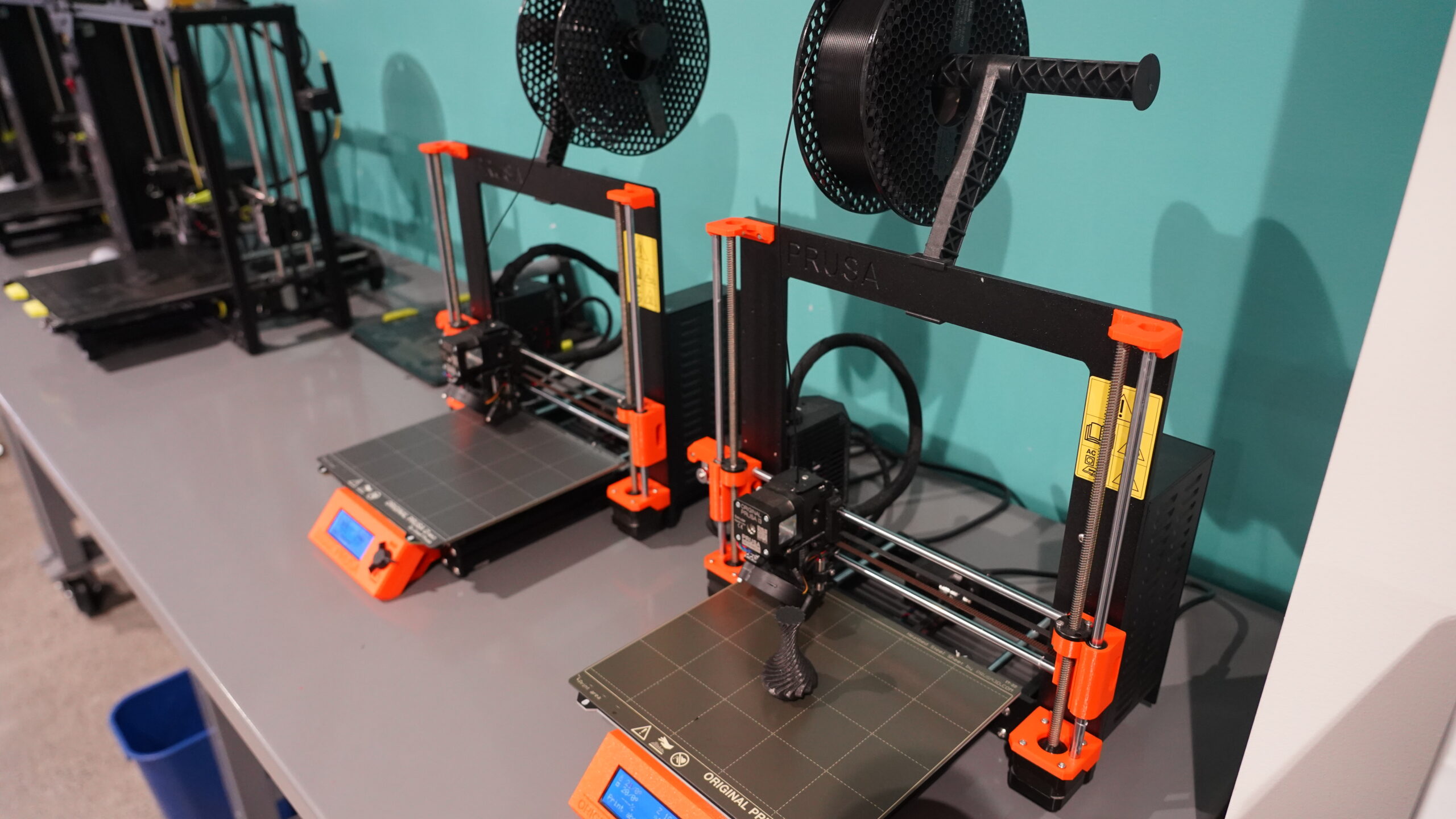 Three 3D printers on a table. Small printed vase is shown on one print bed surface.