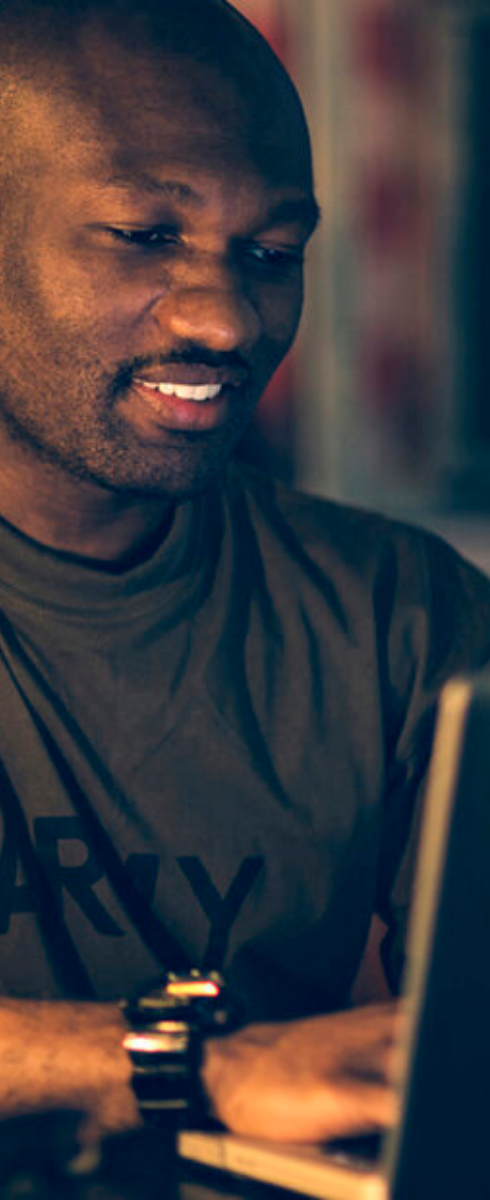 Person in an Army T-shirt using a laptop, with an American flag in the background and a circular badge reading "Veterans & Military Affiliated Students" featuring military branch logos and the WSU logo.