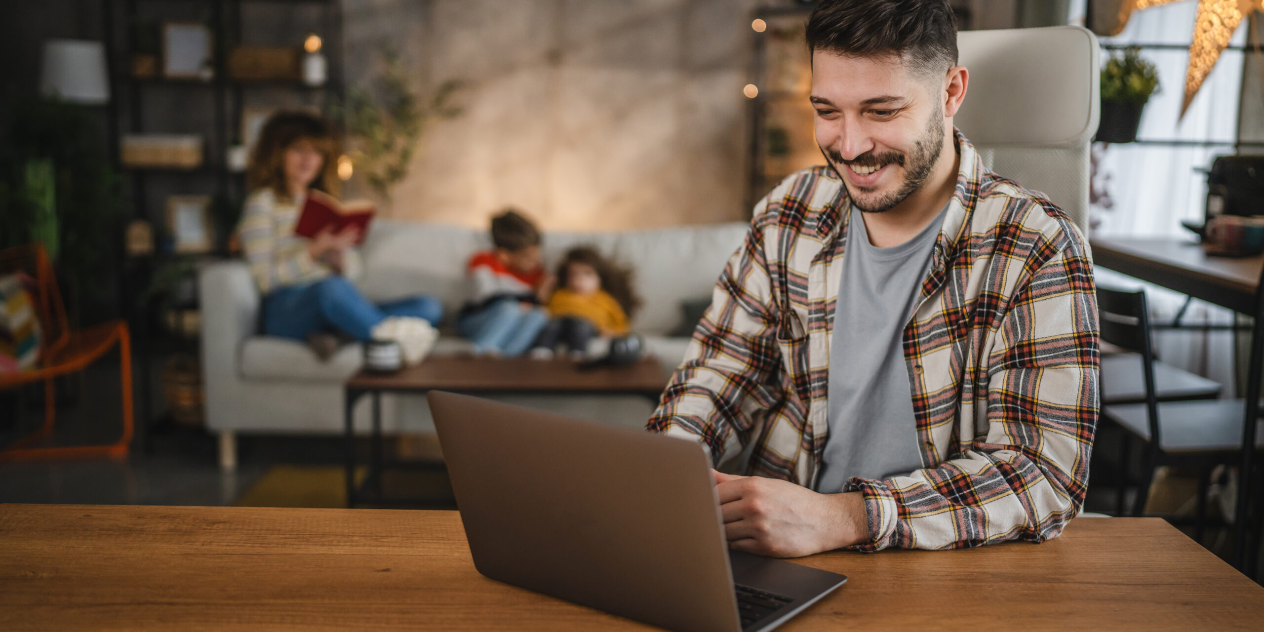 A young man sits at a wooden desk working on a laptop, smiling as he looks at the screen. In the background, a woman is reading a book on the couch while two children sit nearby. The room is warmly lit and decorated with shelves, plants, and cozy furniture.