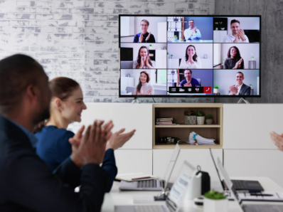 Photo of a meeting room with people applauding and other people on a video call.