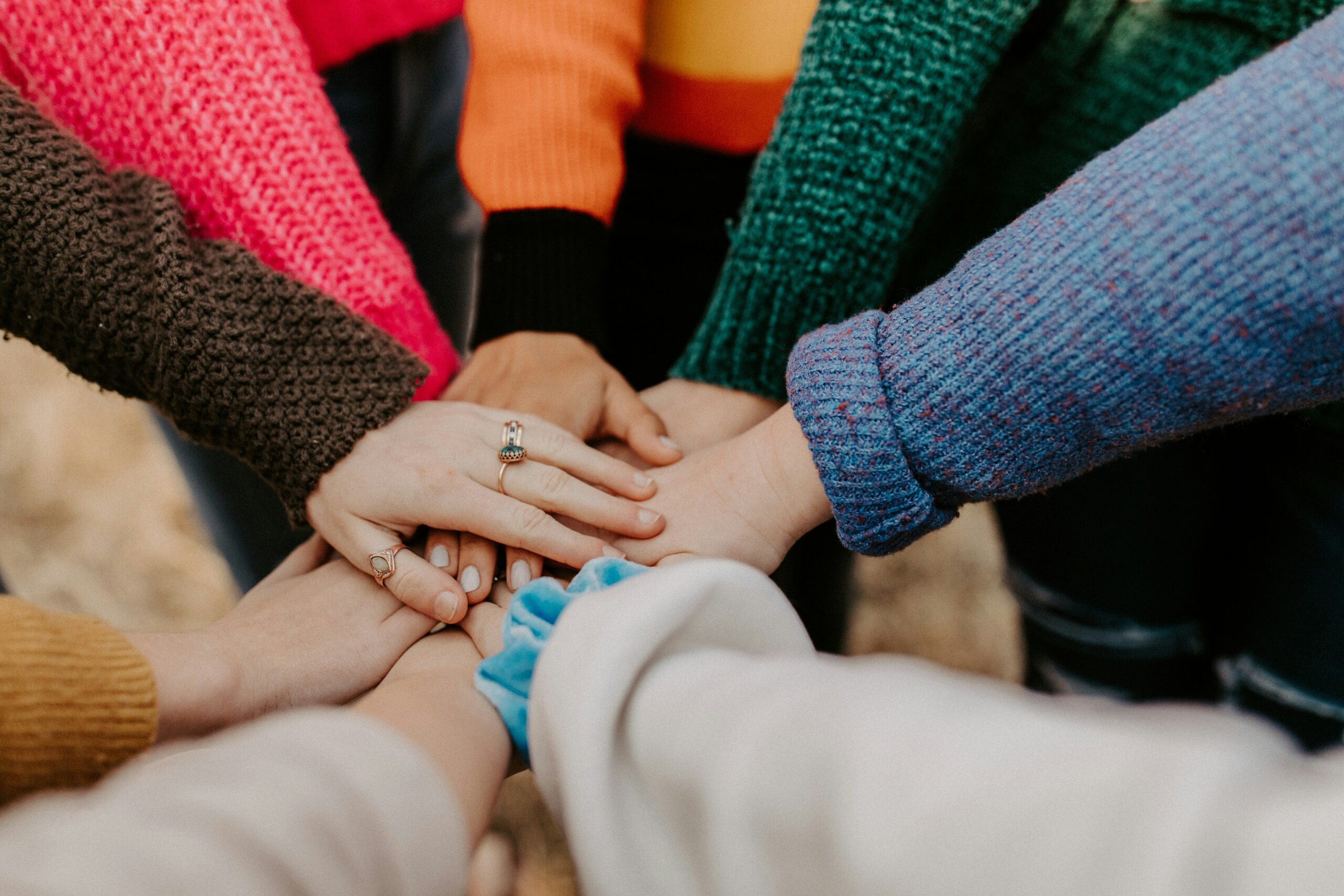 7 hands stacked on top of each other in a circle, with the people wearing brightly colored clothing.