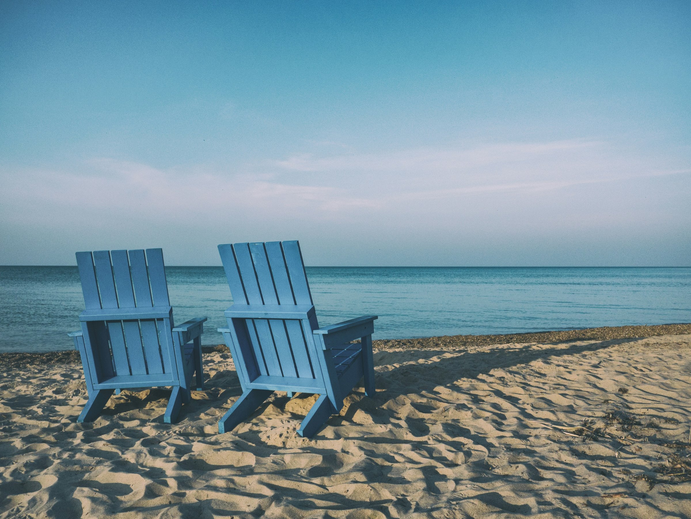 Two blue Adirondack style chairs facing the ocean on a sandy beach.
