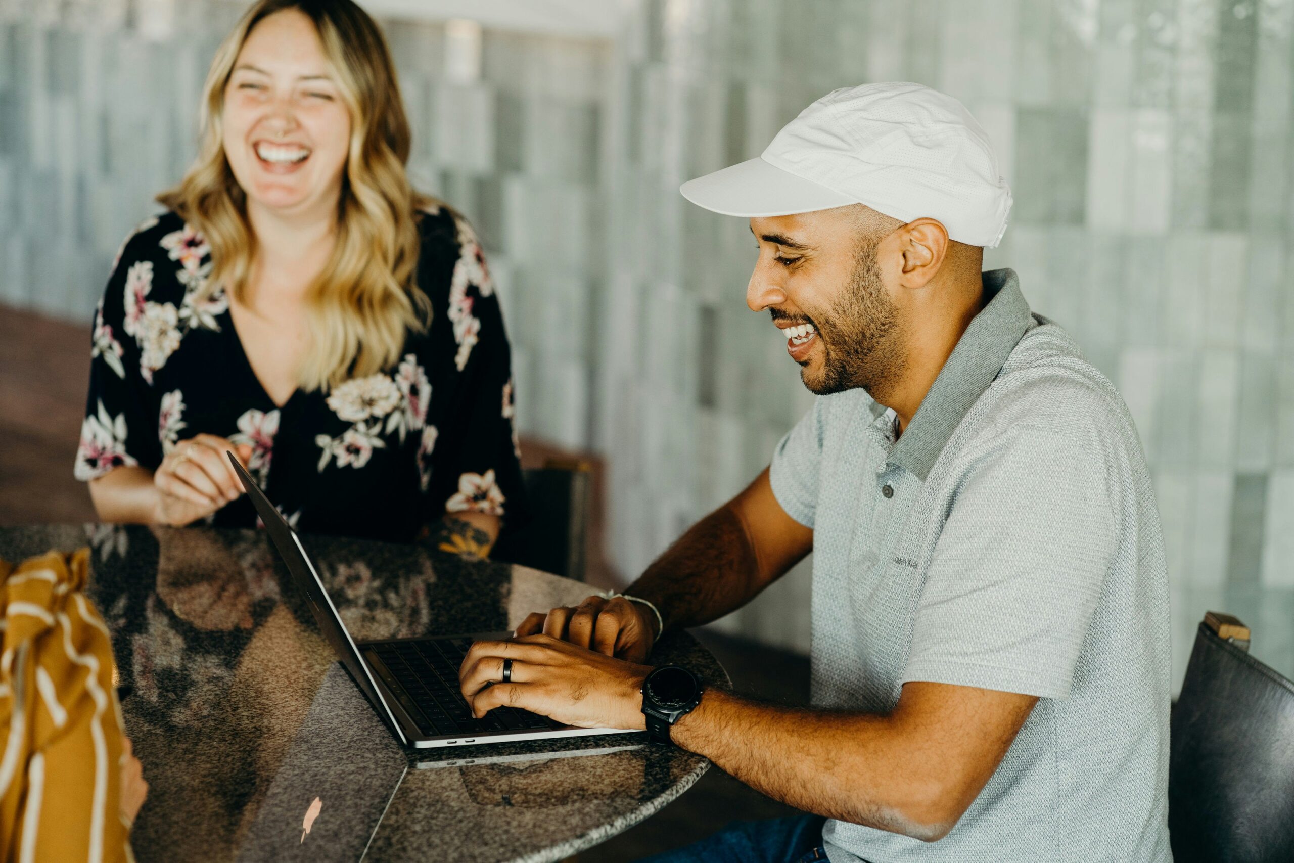 A man in a pale shirt and a woman in a dark shirt, sitting around a table with a laptop laughing and working together.