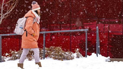 A woman wearing winter coat and hat, walking in the snow in front of a red glass building.