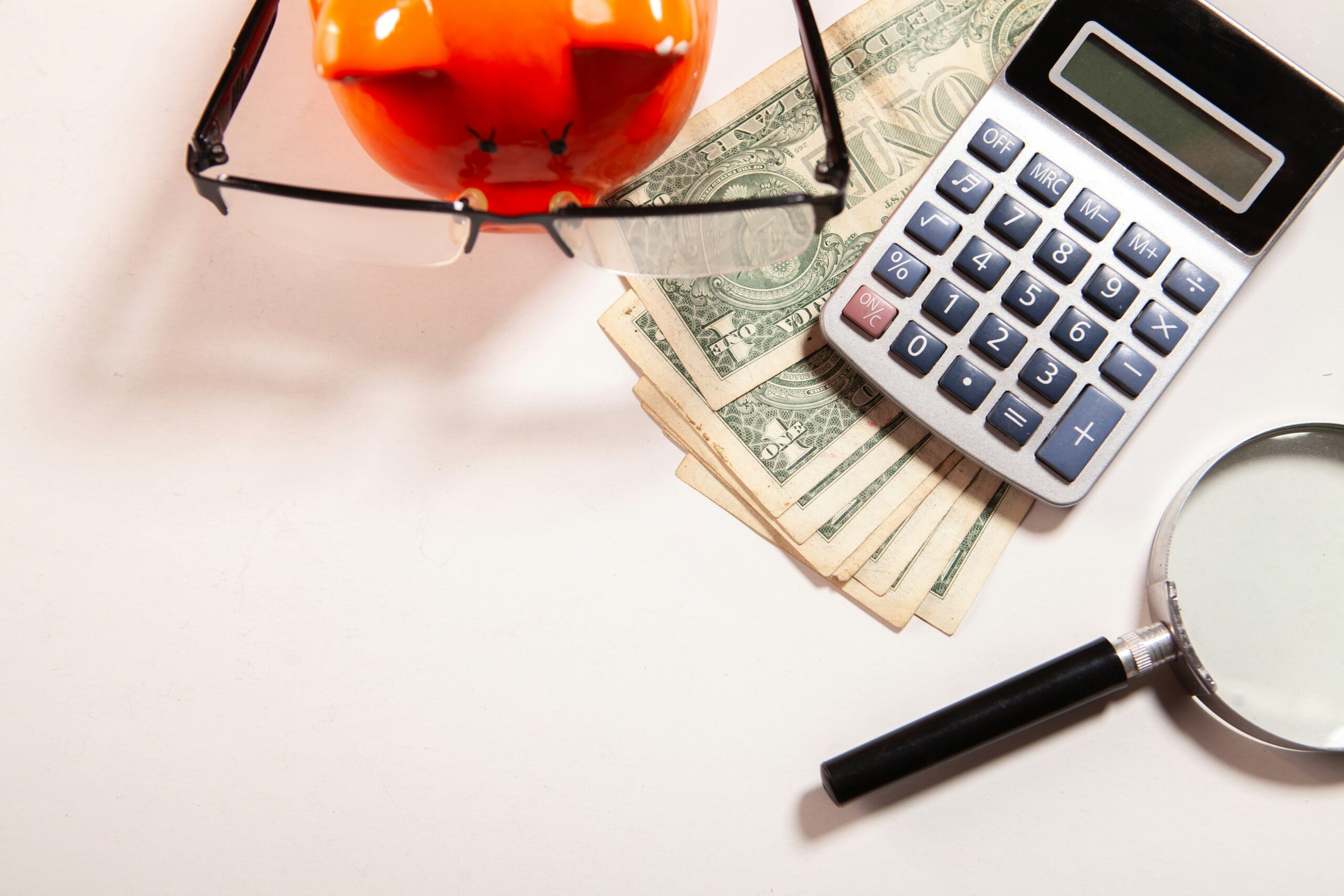An arrangement of a magnifying glass, a calculator, a pile of 1 dollar bills, and an orange piggy bank wearing glasses.