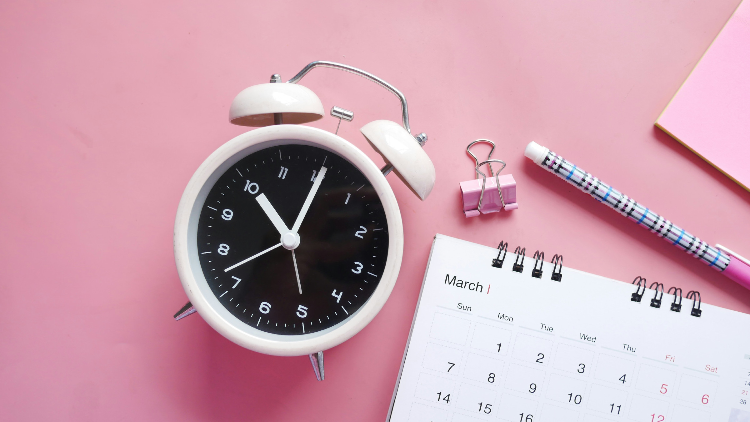 A white alarm clock and a calendar on a pink background.