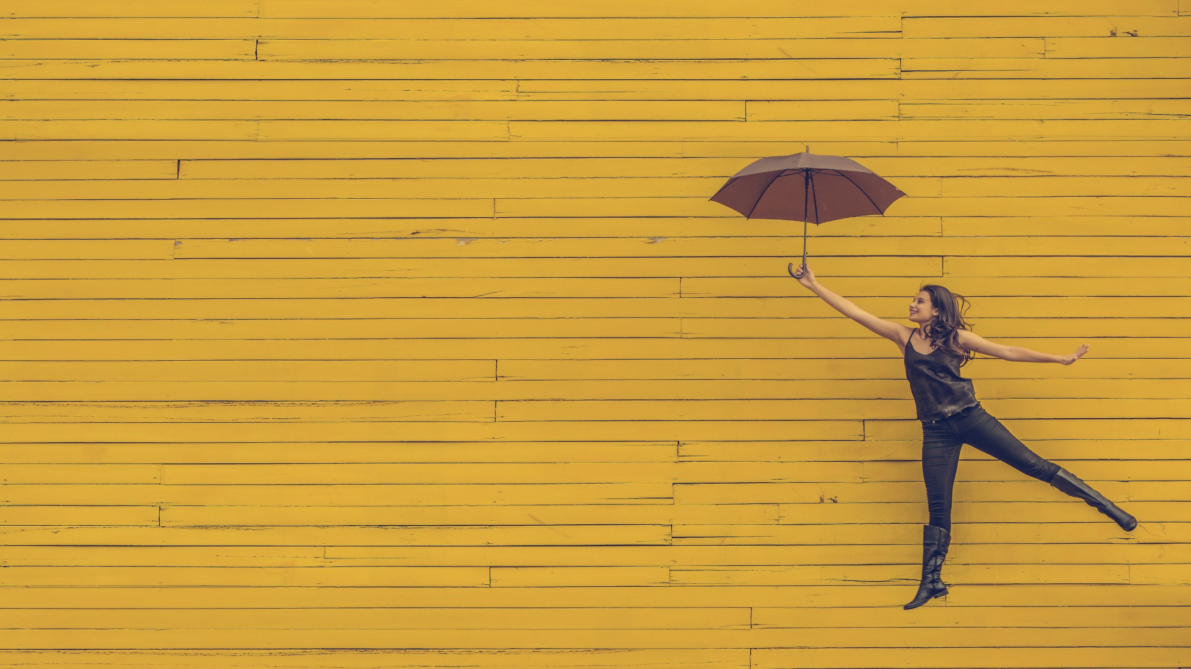Woman in brown, holding an umbrella, jumping in front of a wooden, yellow wall.