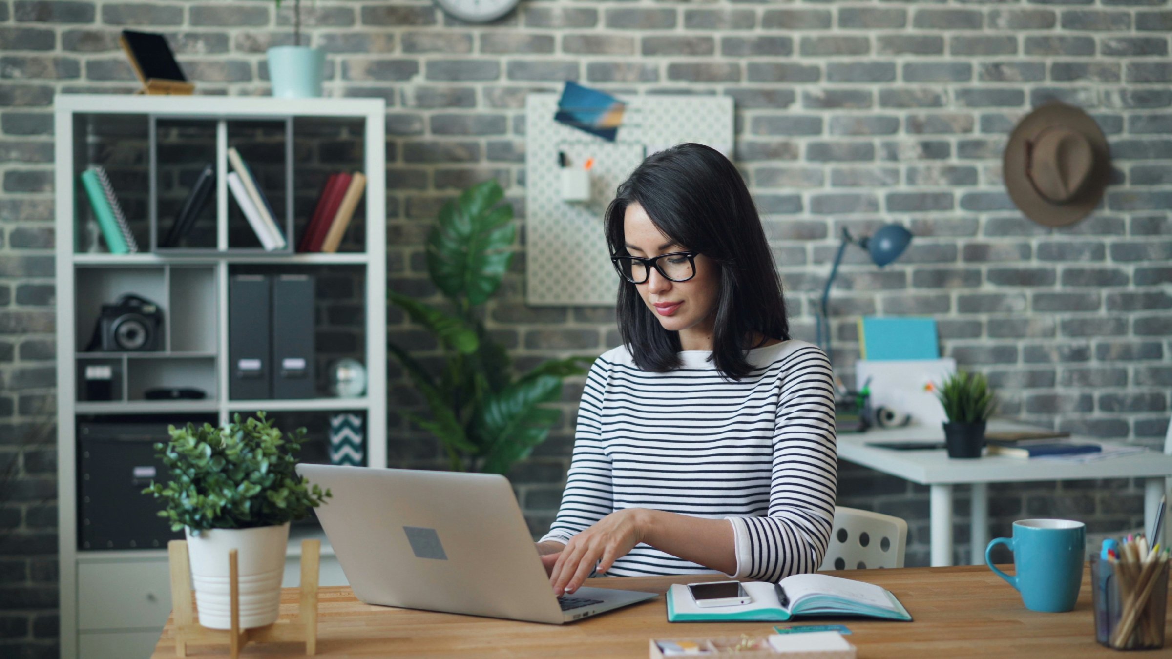 A woman with dark hair and wearing glasses sitting at a desk working on a laptop.