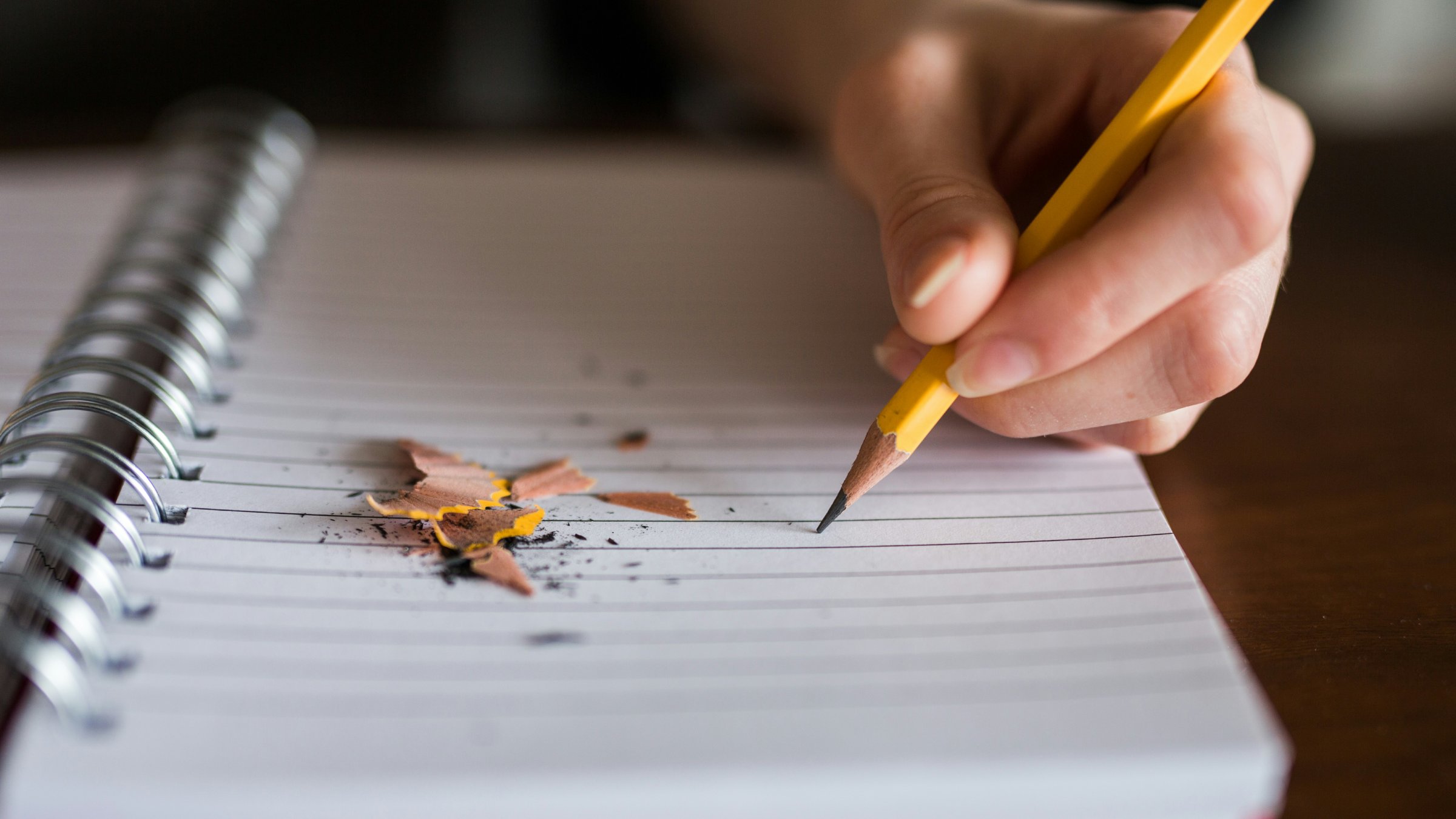 A hand holding a pencil on a blank page, ready to write, with the tip of the pencil surrounded by pencil shavings.