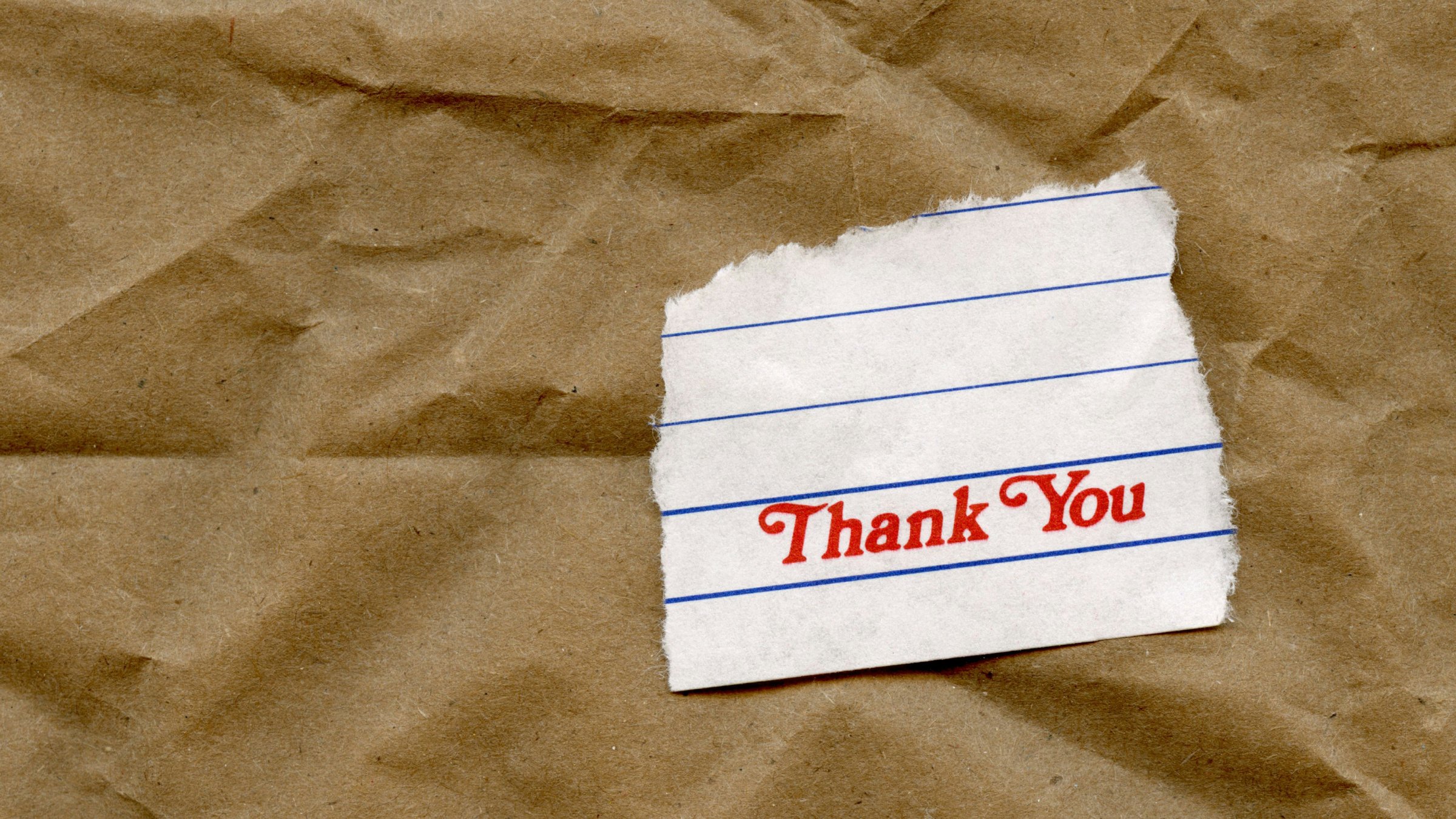 A torn off sheet from a note pad laying on a brown paper bag, with the word "Thank You" printed on the bottom of the sheet.