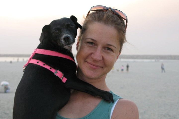 Kristi holding a small black dog. A body of water is behind them.