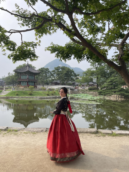 WSU student in a hanbok by a pond with a Korean pavilion and mountains in the background.