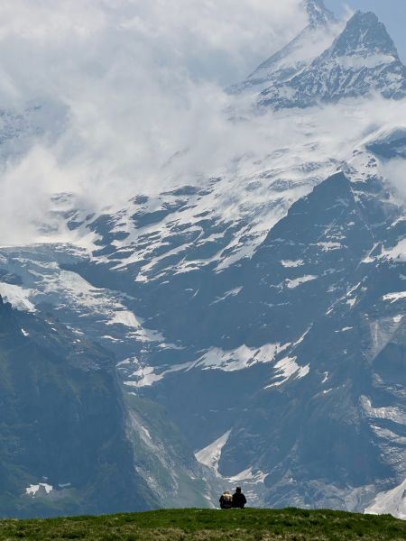 A mountainous landscape with snow-capped peaks and two people sitting in the foreground.