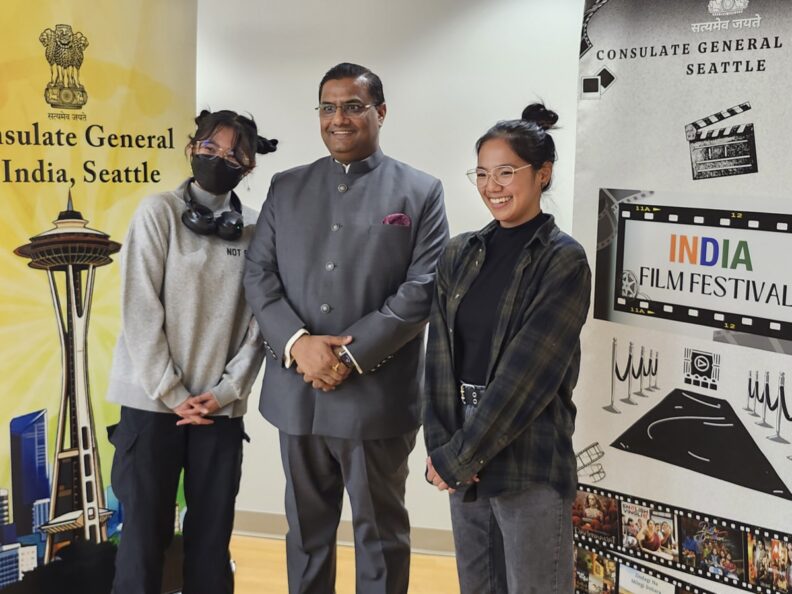 Three people stand indoors between two vertical banners. The banner on the left reads “Consulate General of India, Seattle” and features a stylized Seattle skyline. The banner on the right reads “India Film Festival” with film-related graphics. All three people are facing the camera; one wears a gray suit, and the other two wear casual clothing.
