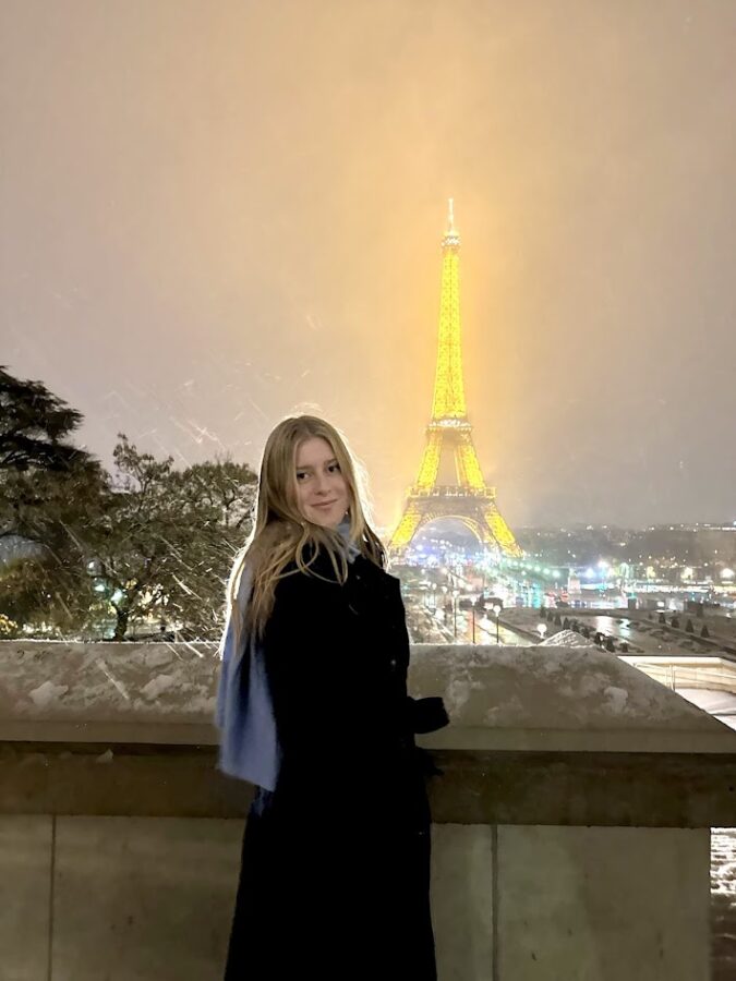 A person stands at a stone railing at night with the illuminated Eiffel Tower glowing yellow in the background as light snow falls over the city of Paris.