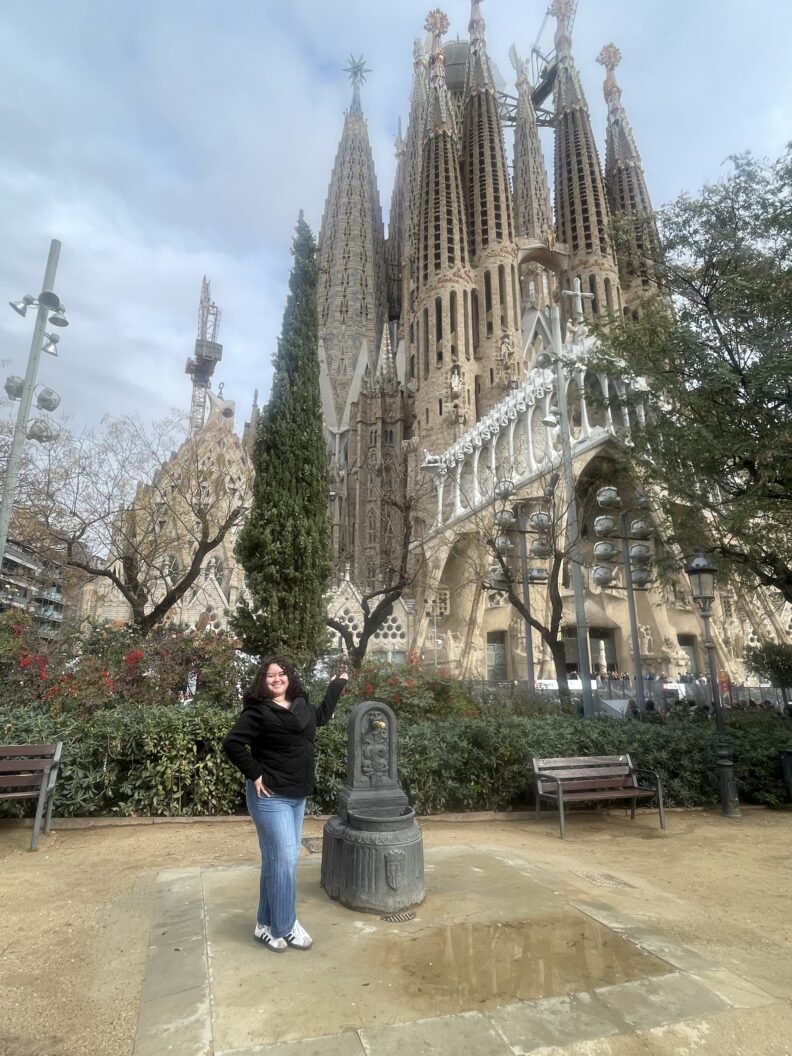 A person stands on a paved area in a small park with benches and trees, posing beside a short stone sculpture, with the tall, ornate towers of the Sagrada Família in Barcelona, Spain, rising in the background.
