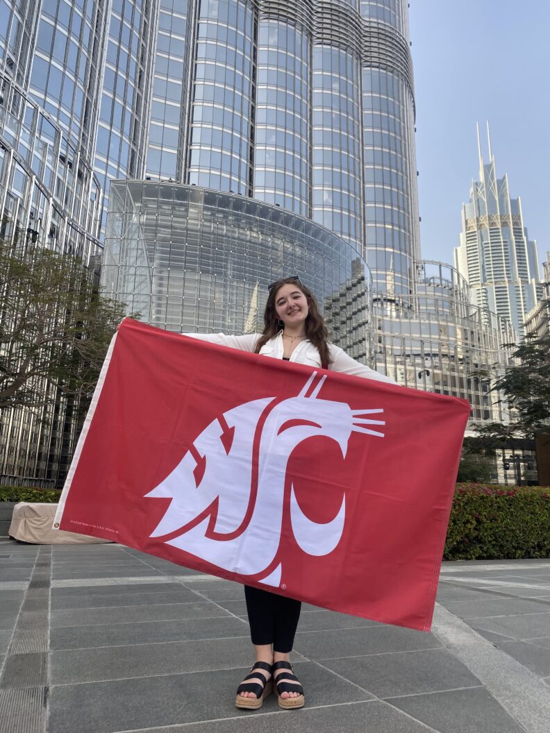 A person stands in an outdoor plaza holding a red Washington State University flag, with modern glass skyscrapers surrounding the space in the background.