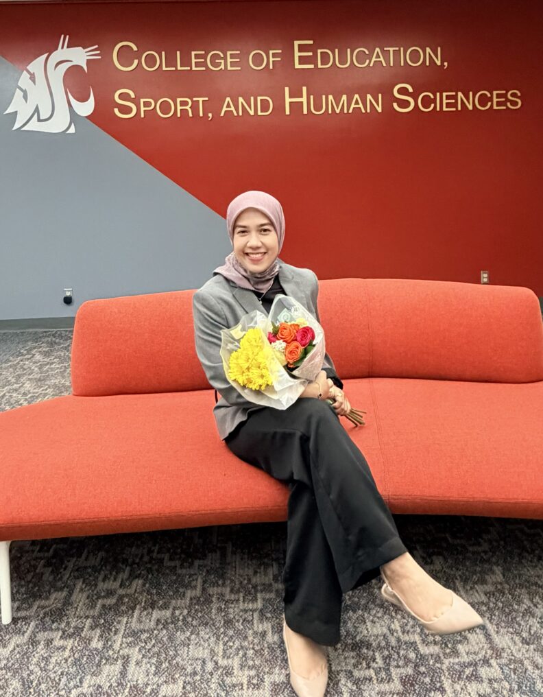 A person sits on a red couch holding a bouquet of flowers, with a wall behind them displaying the text “College of Education, Sport, and Human Sciences” and a Washington State University logo.