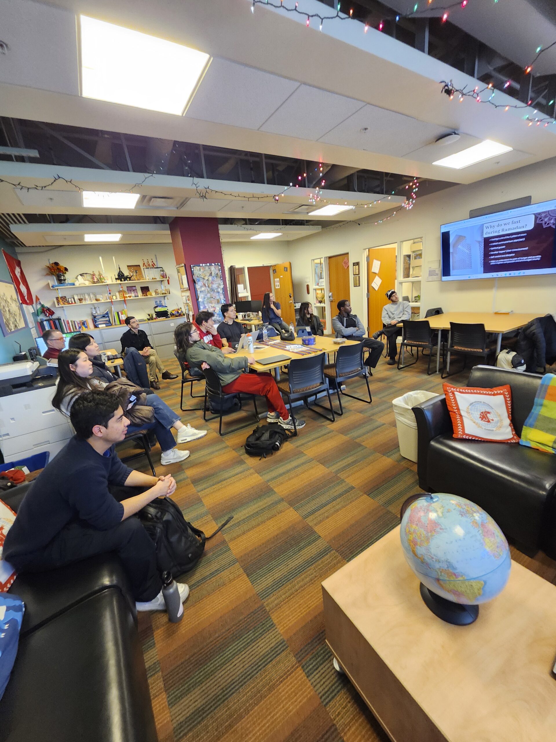 About 12 people in a meeting room watch a presentation slide about fasting during Ramadan on a wall-mounted screen.