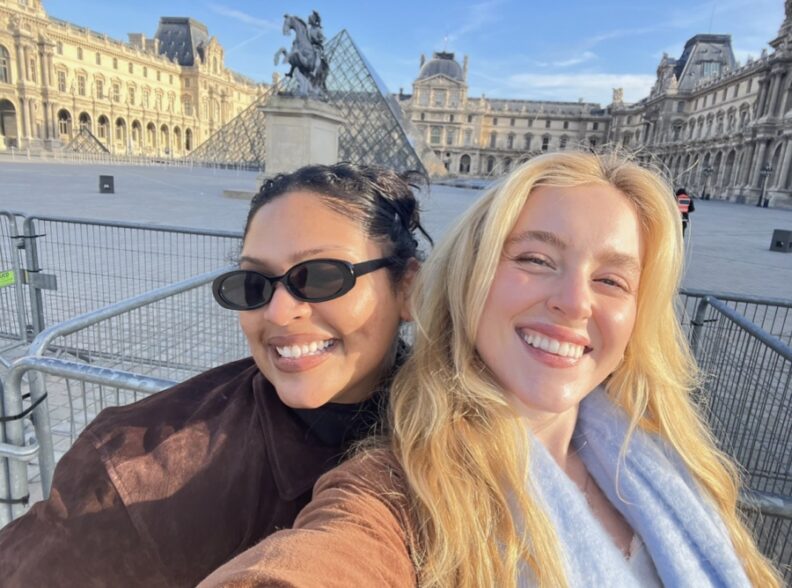 Two people take a selfie in the courtyard of the Louvre Museum in Paris. The Louvre’s glass pyramid and historic palace buildings are visible in the background, with metal crowd-control barriers surrounding the courtyard under a clear blue sky.