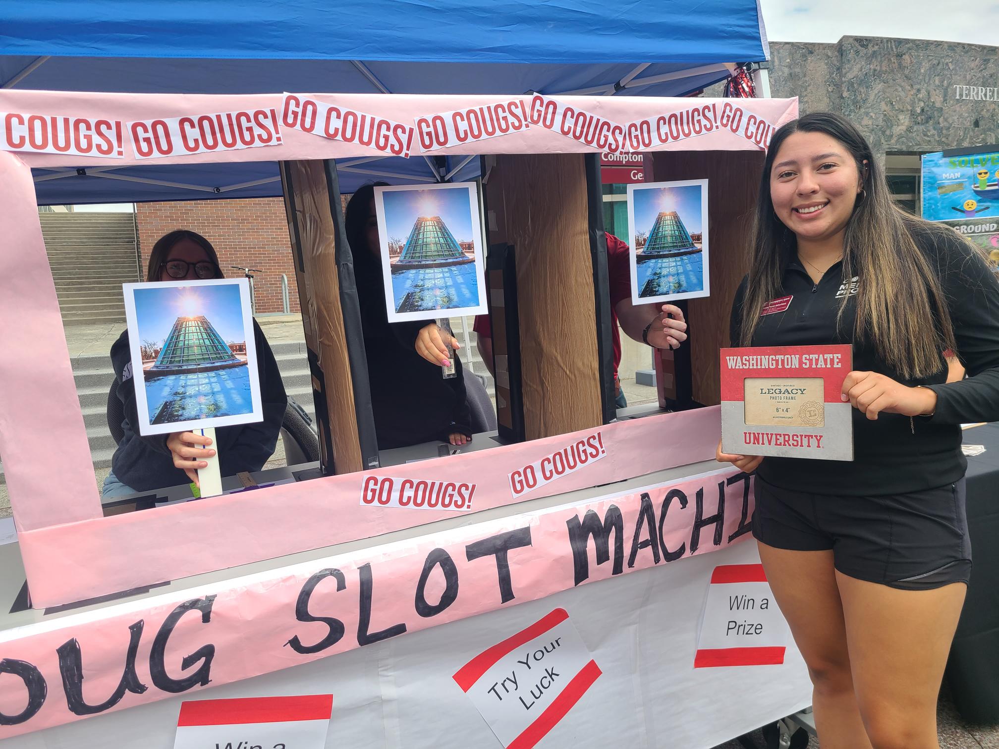 WSU Picnic Highlights WSU student at “Coug Slot Machine” booth holding a Legacy certificate, with “Go Cougs!” signs and prizes displayed.
