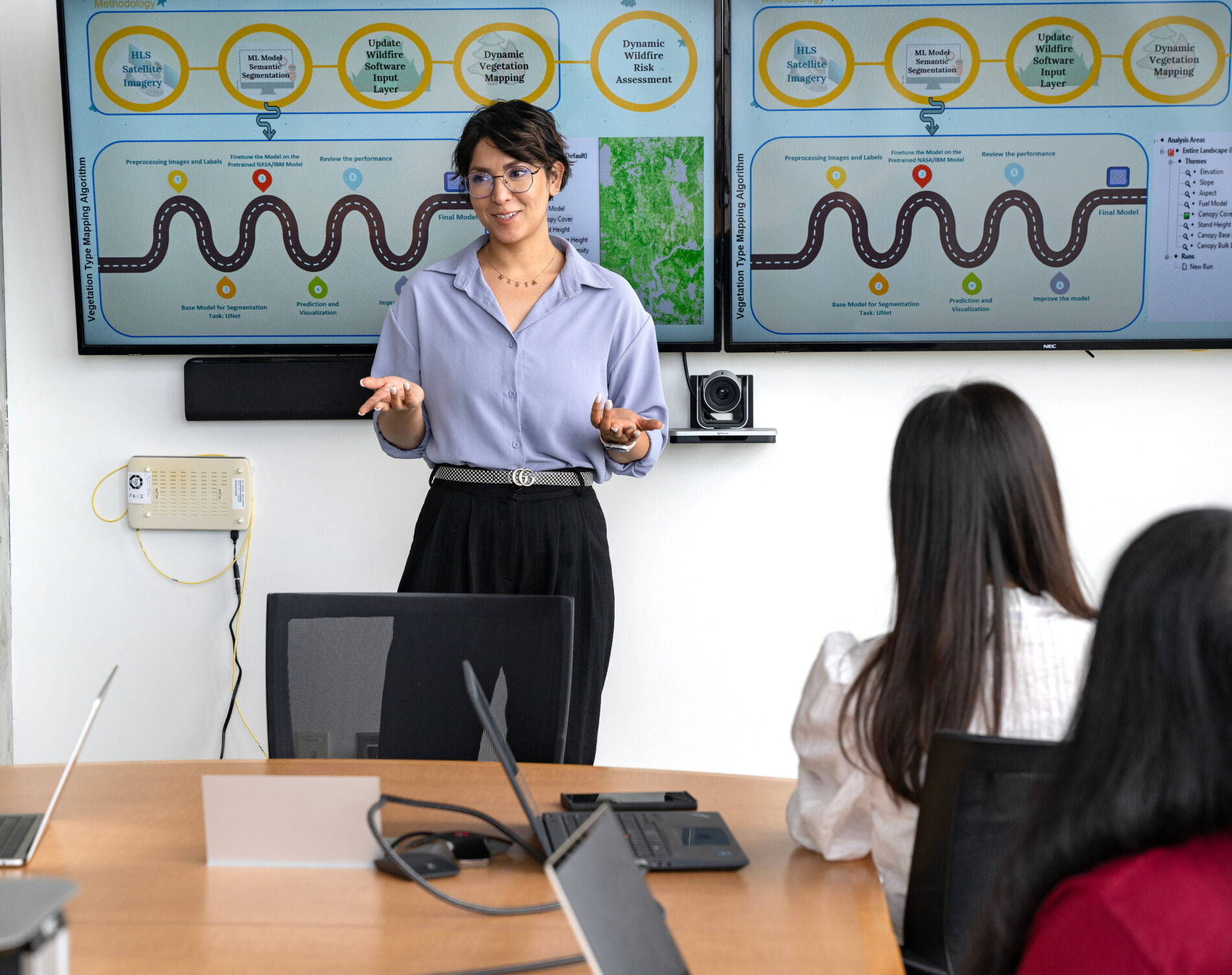 Three researchers in white lab coats work together in a red-lit laboratory, reviewing data on a computer screen and recording notes on a clipboard.