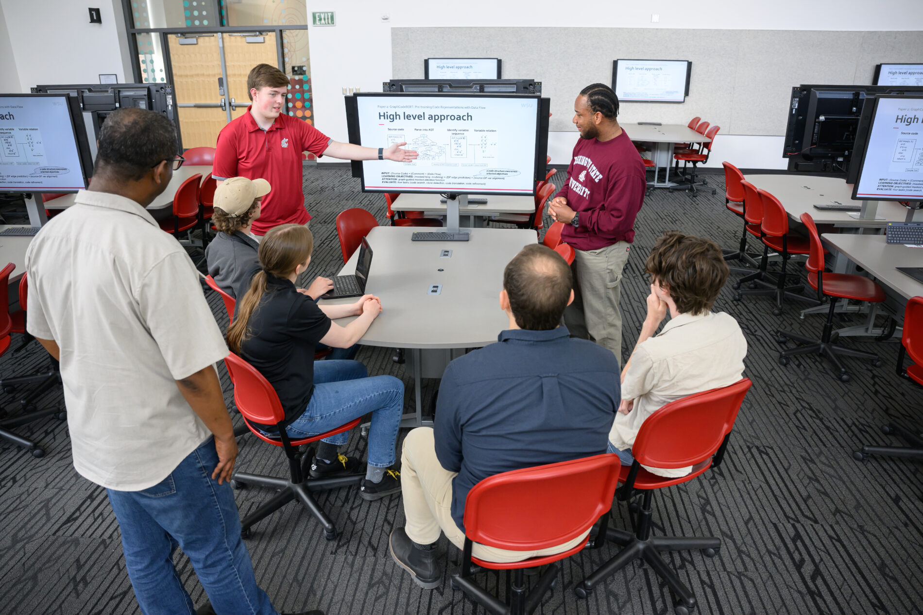 A group of students seated around a table attentively listen as two instructors stand and present a technical diagram on a large screen labeled 'High level approach' in a modern classroom with multiple monitors and red chairs.
