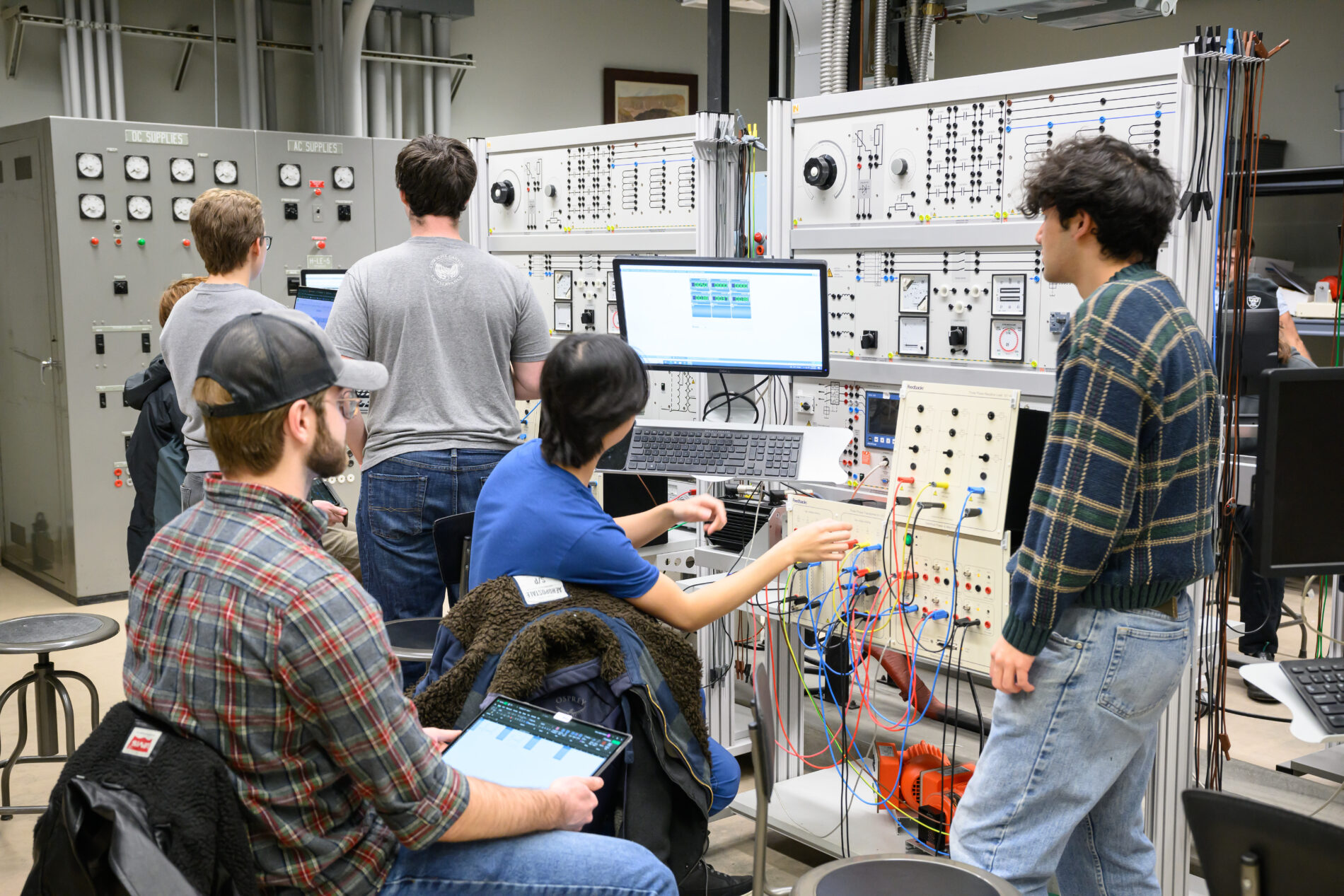 Engineering students working in a lab with control panels, wiring boards, and computer monitors while conducting hands-on experiments with electrical circuits.