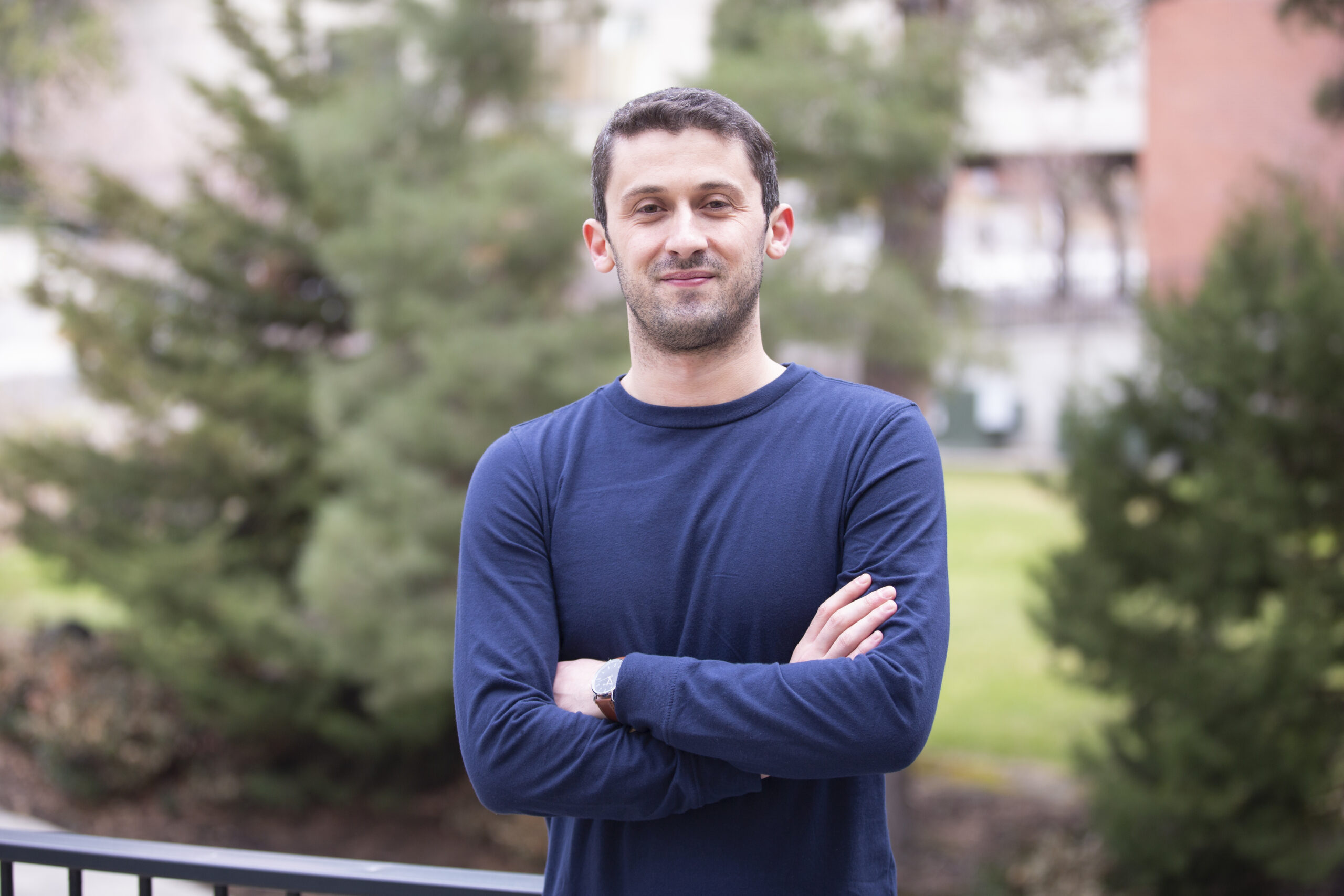 A portrait of Onur Ramazan standing outside next to trees and a brick building.
