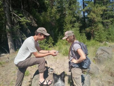 Two people stand next to each other looking at a rock. A dog watches. 