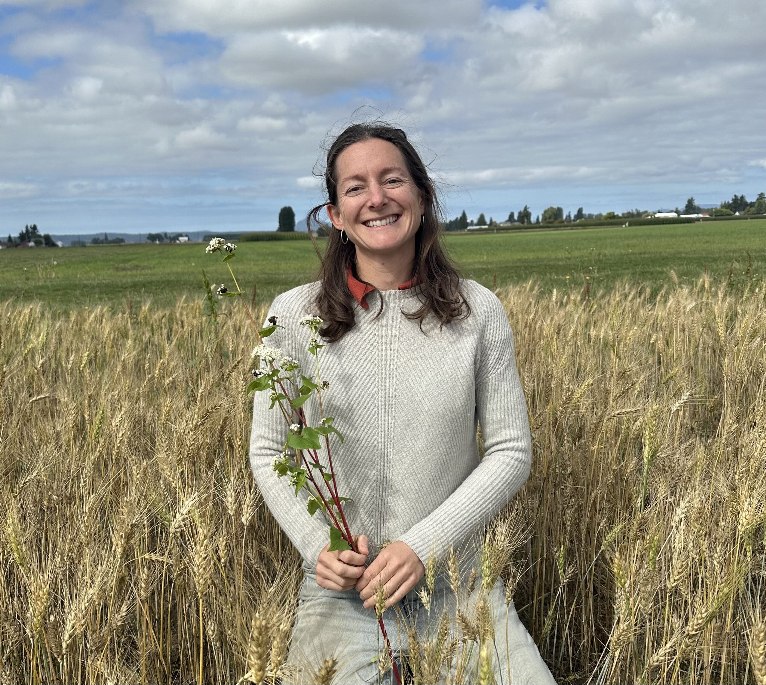 Annah smiling in a barley field while holding a buckwheat plant.