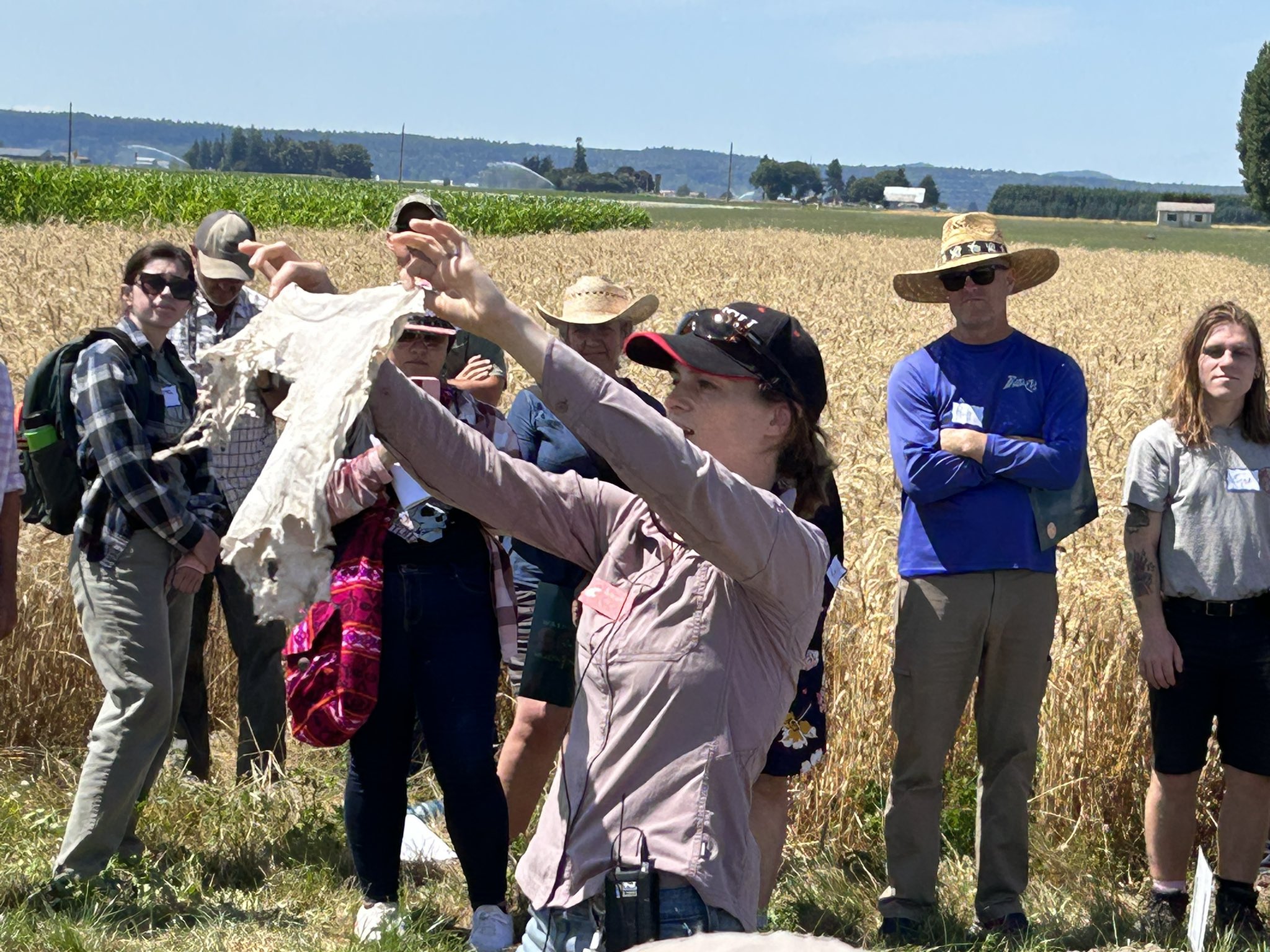 Deirdre Griffin LaHue stands in front of a group of people by a wheat field, holding up a microbially degraded shirt.