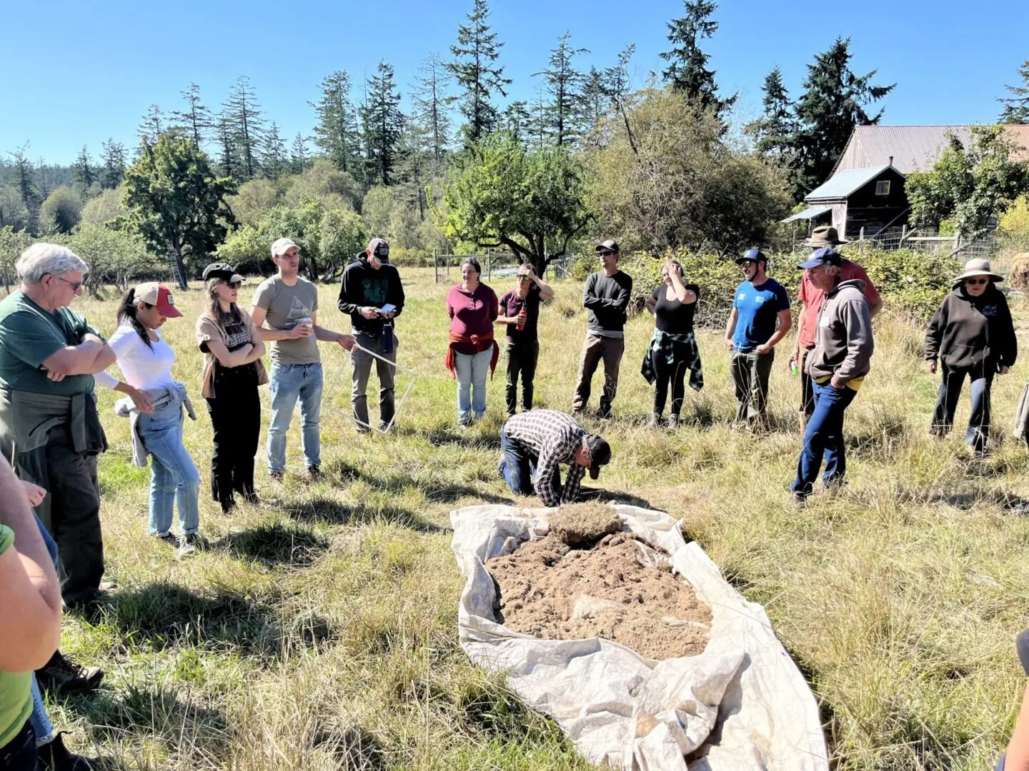 Gabe LaHue presents to a group of farmers gathered around a cloth tarp filled with soil, while one farmer kneels to closely inspect it.