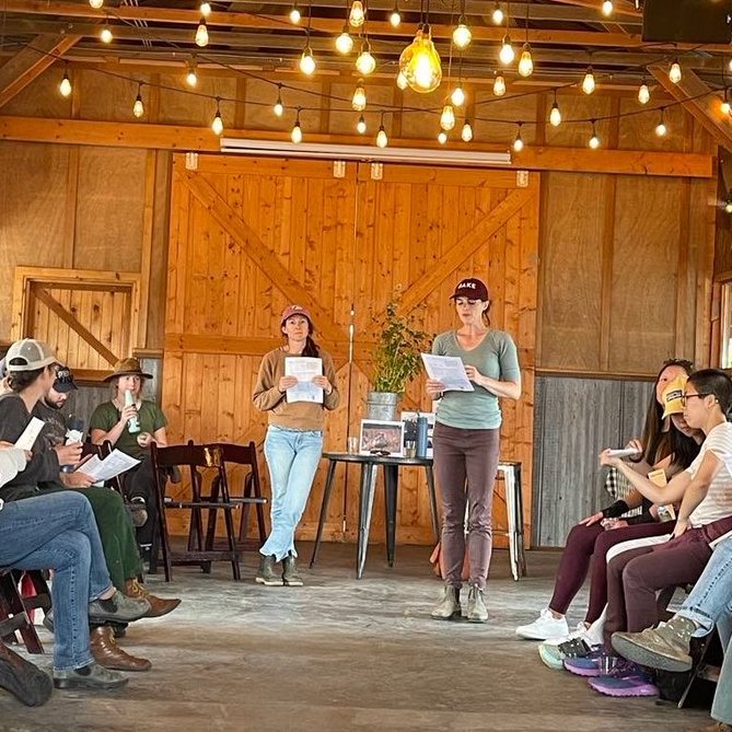 Deirdre and Annah in a barn, presenting to an audience, with attendees seated along the sides of the room.