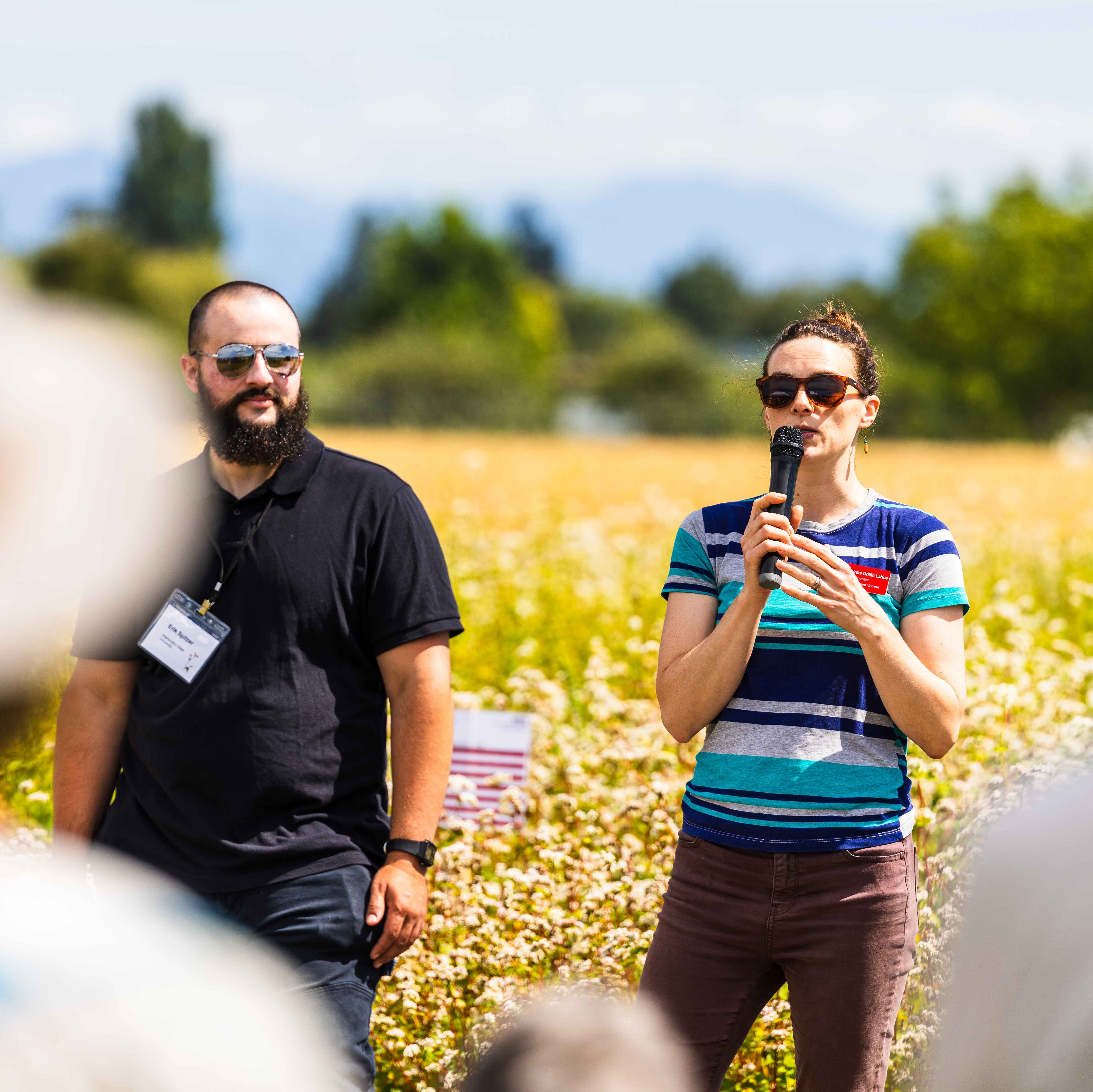 Deirdre Griffin LaHue and PhD student Erik Spitzer presenting at a field day, with a buckwheat field behind them; Deirdre is holding a microphone.