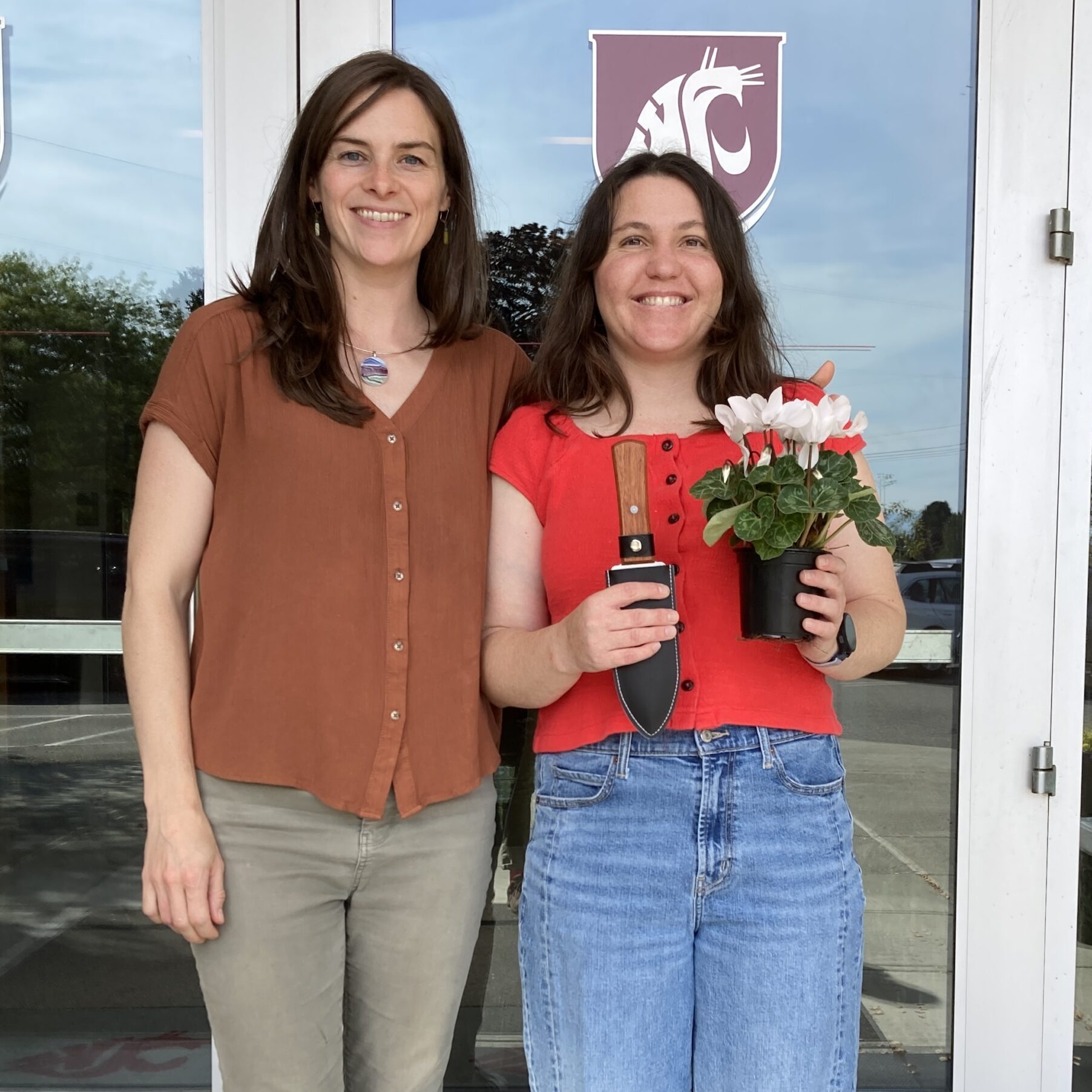 Deirdre and Madeline smiling and standing in front of a door with a WSU logo; Madeline holds a potted plant and soil knife.