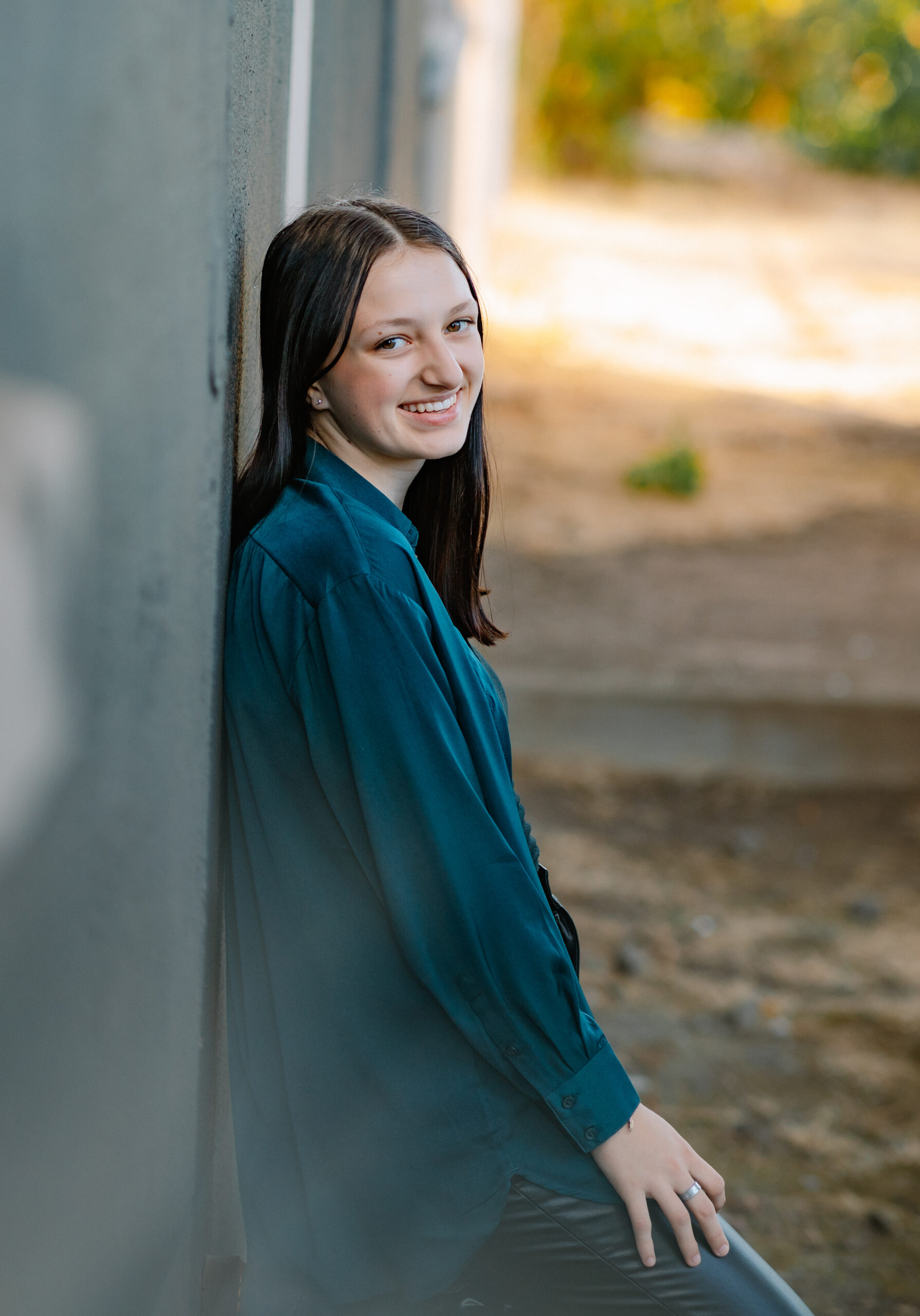 A woman with brown hair and a blue shirt leaning against a wall and smiling.