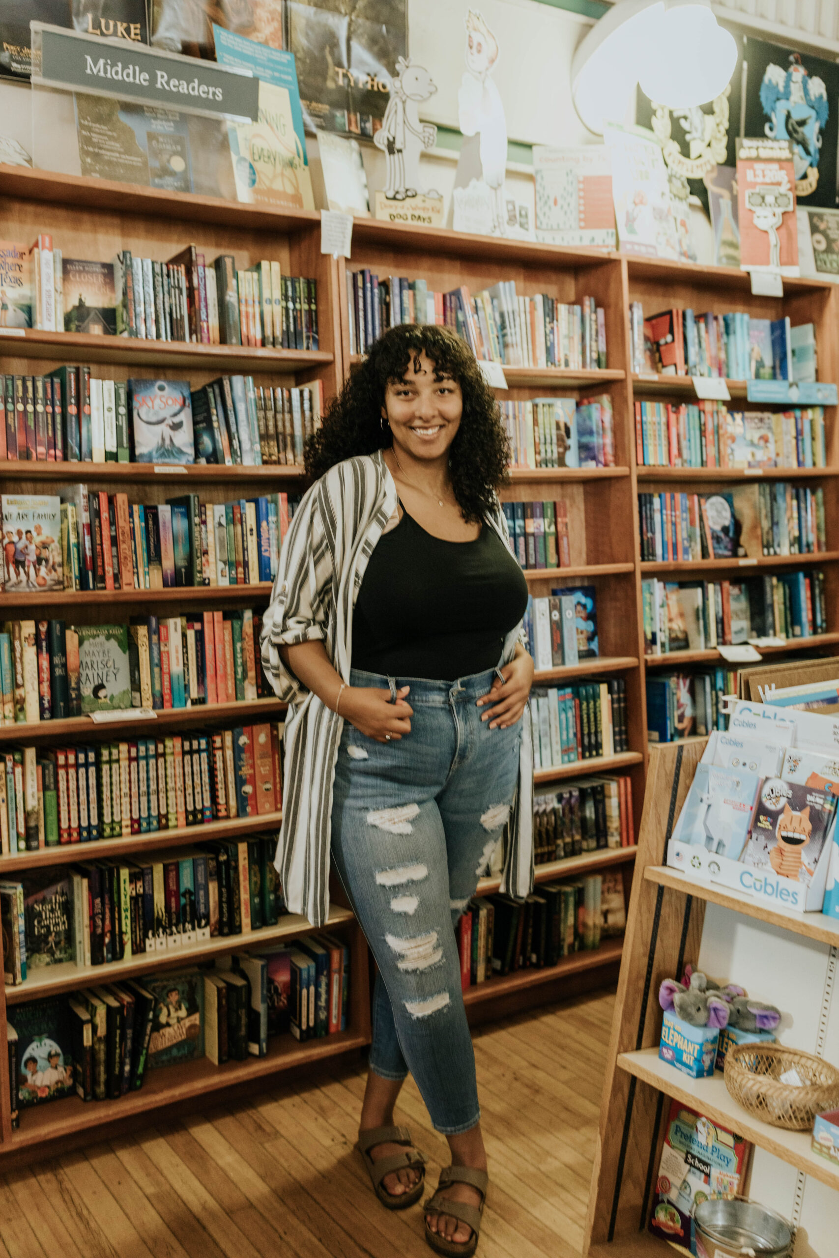 A woman with black curly hair standing in front of a set of bookshelves.