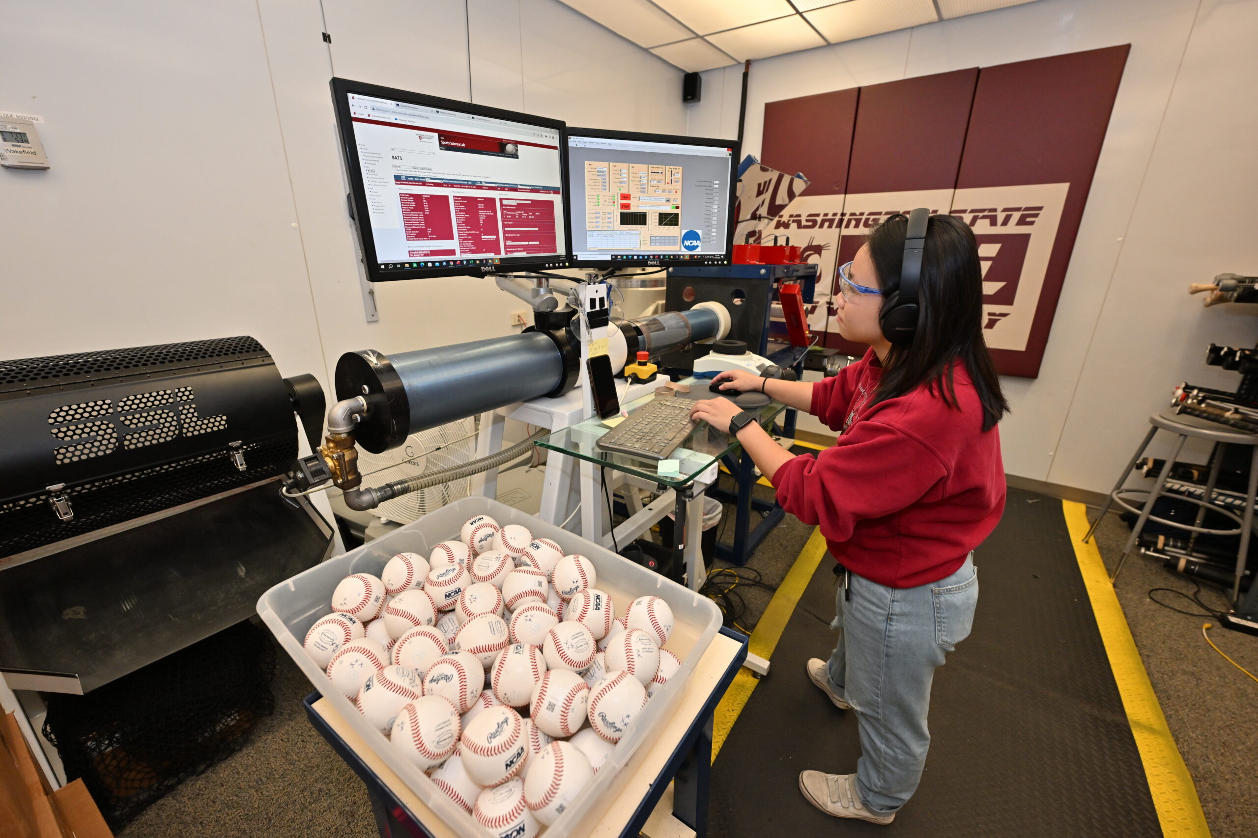 WSU Grad Student testing a bat in the Sports Science Lab. A student is standing in front of a computer console wearing ear protection. Next to the computer console is a tub of baseballs and behind the computer is a canon that launches baseballs.