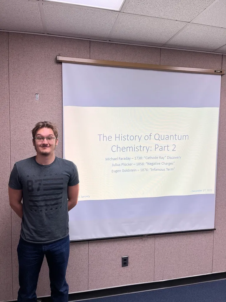 Jonah Sprosty giving his presentation. The screen reads: The History of Quantum Chemistry: Part 2. Michael Faraday - 1738: "Cathode Ray" Discovery. Julius Plucker - 1958: "Negative Charages". Eugen Goldstein - 1876: "Infamous Term".
