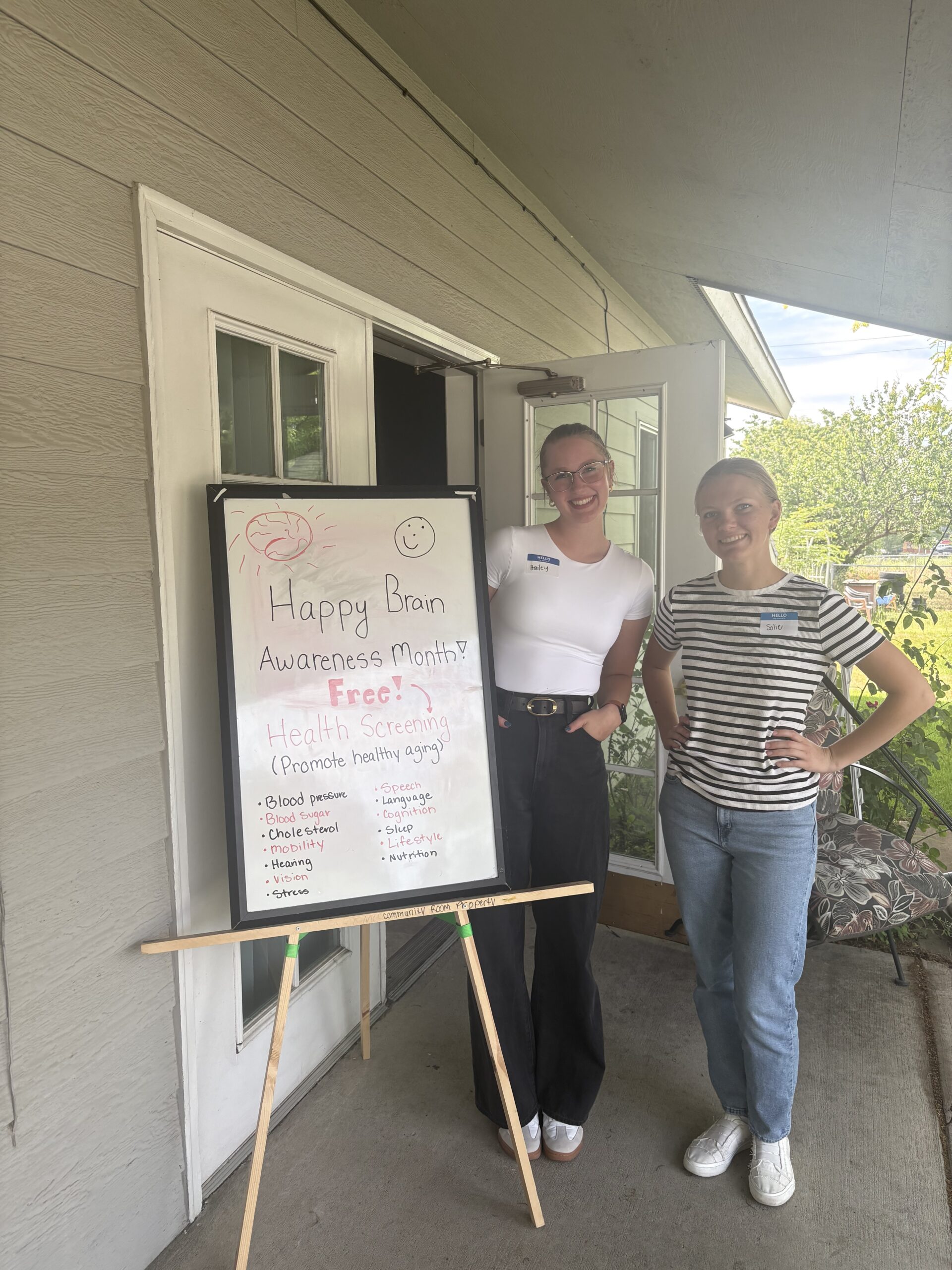 Two lab members outside a BRAIN-FIT event with a sign that promotes the event.